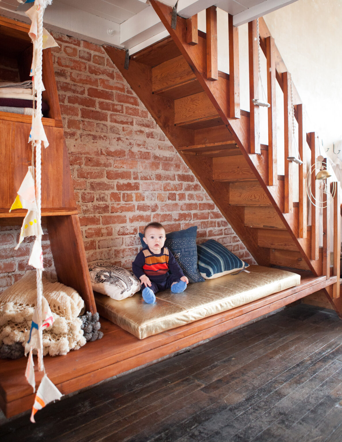 Baby sitting up in a nook under a stairway made of a wooden floating bench and a metallic gold banquette pillow